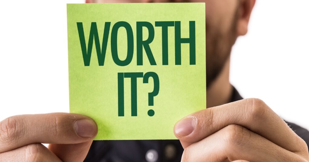 A close-up of a person holding a green sticky note with the words 'WORTH IT?' written in bold, dark green text. The note is centered in the image, with the person's face slightly blurred in the background, creating a focus on the question.