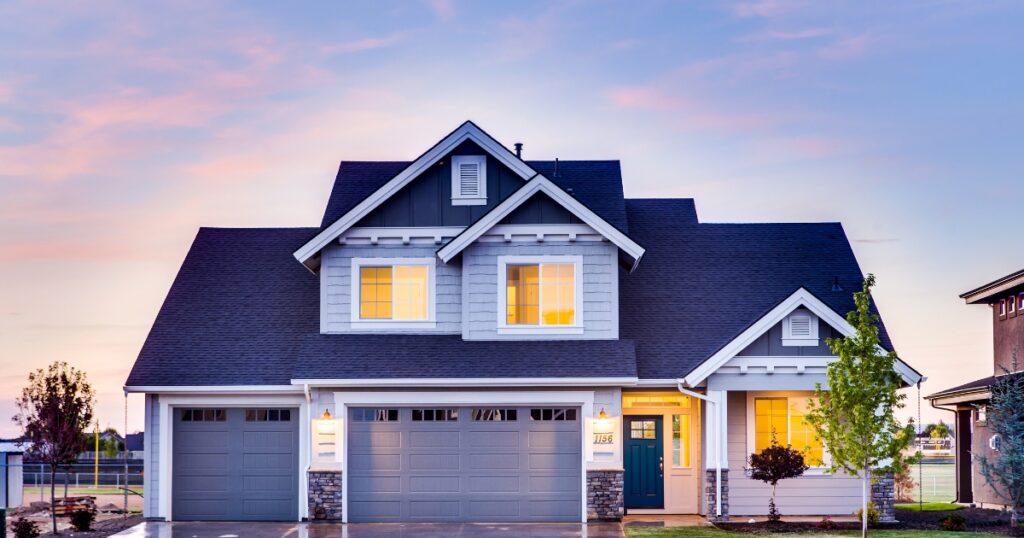 A modern two-story house with a grey and white exterior, a dark blue roof, and three illuminated garage doors. The front door, painted a deep teal, is surrounded by glowing windows, creating a warm and inviting ambiance. The house is set against a colorful sunset sky with soft pink and purple hues.