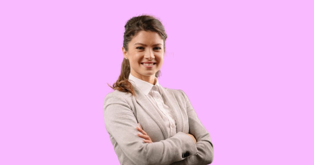 A confident young woman with brown hair tied back, wearing a white collared shirt and a light grey blazer, standing with her arms crossed. She is smiling warmly against a solid light pink background, conveying professionalism and approachability.